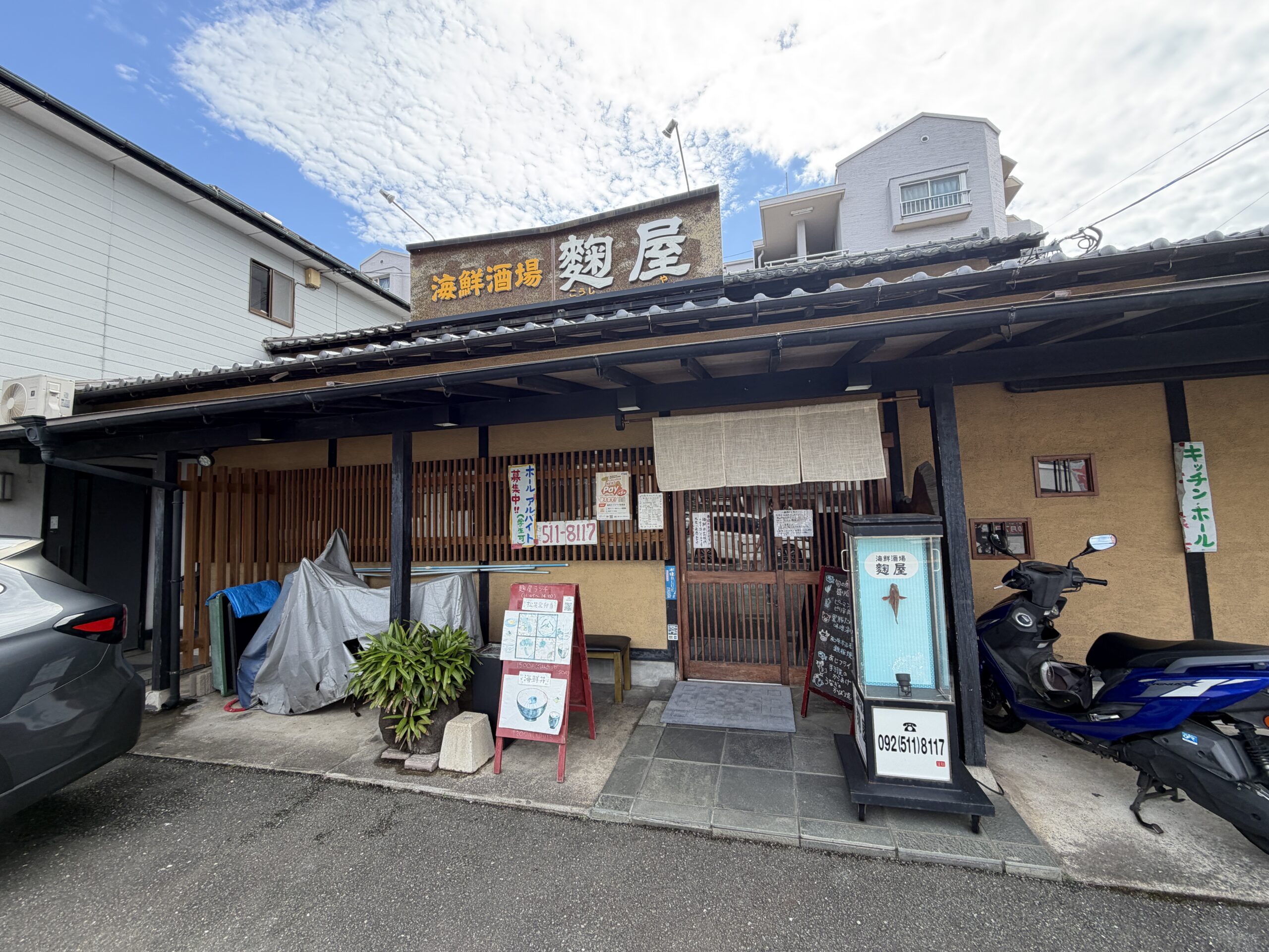 瓦の庇と木の格子、黄土色の壁のお店の外観。木の看板に海鮮酒場 麹屋とある